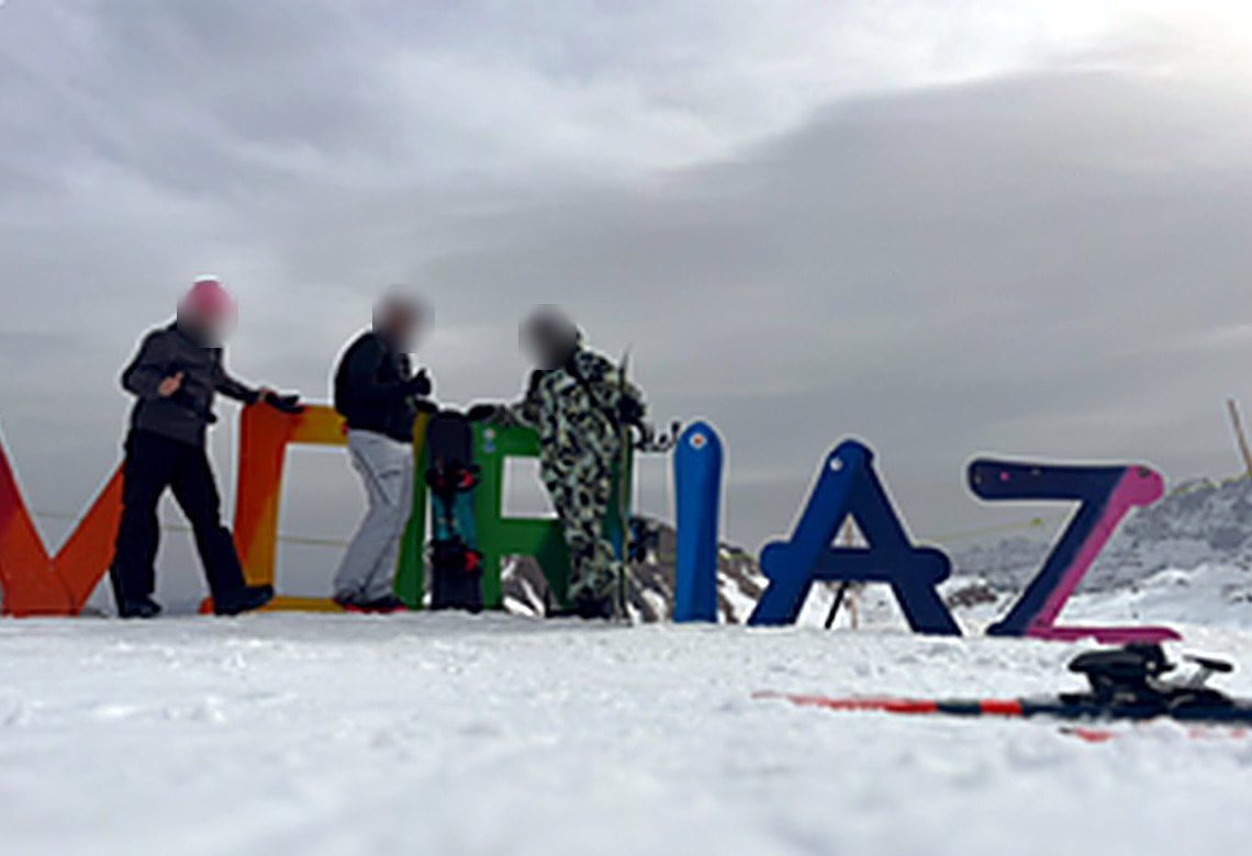 Trois jeunes posent devant les grandes lettres colorées de la station, au sommet des pistes.