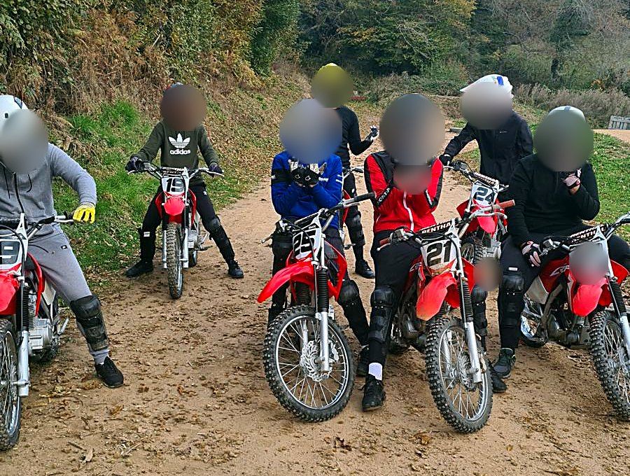 Un groupe de jeunes casqués sur des motos de cross rouges, sur un chemin en forêt, visages floutés.