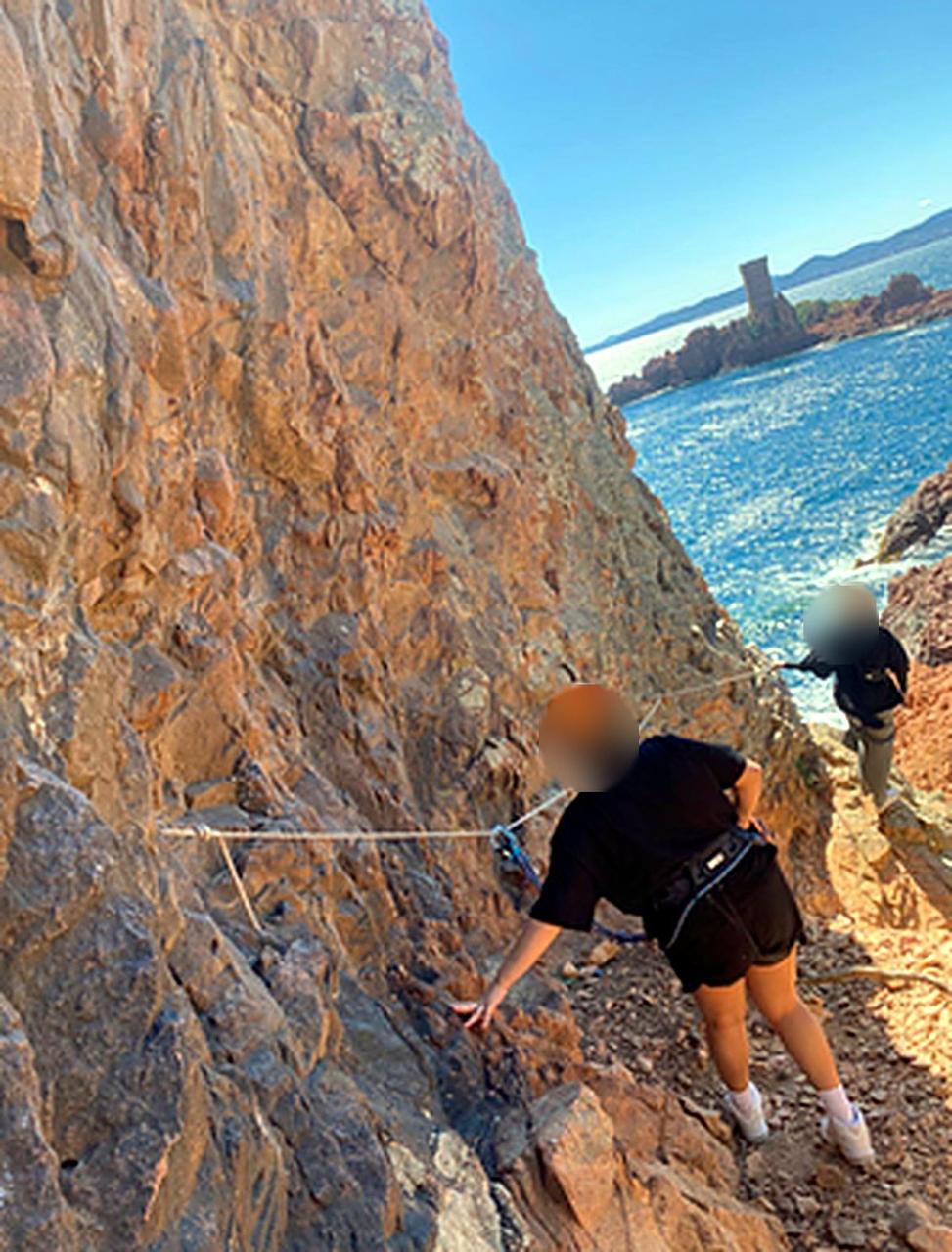Une jeune, casque sur la tête, progresse le long d'une paroi rocheuse au bord de la mer, visages floutés.